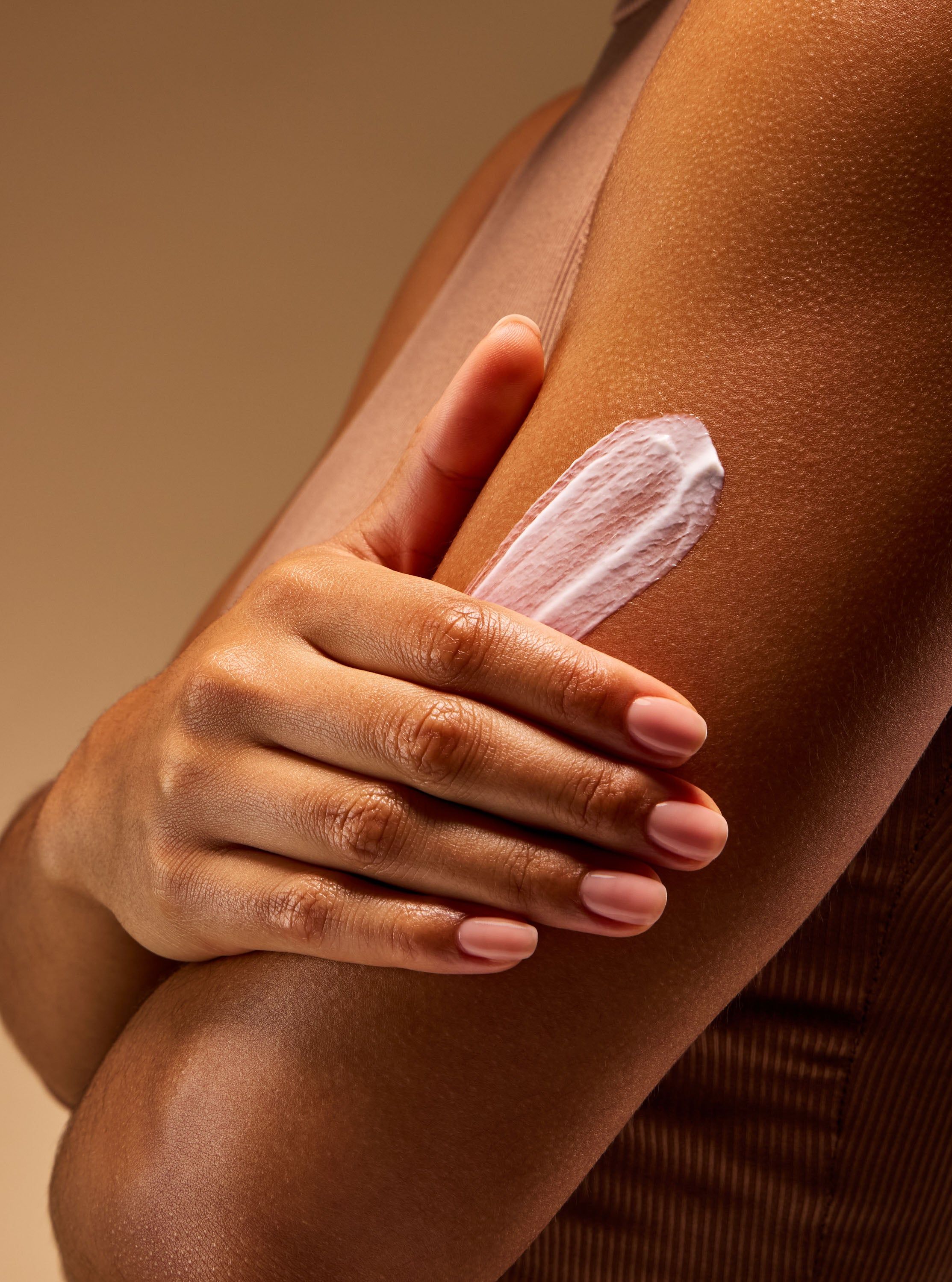 Ruka roztírá krém na pokožce paže, zvýrazňující výživnou, hydratační texturu péče o tělo.