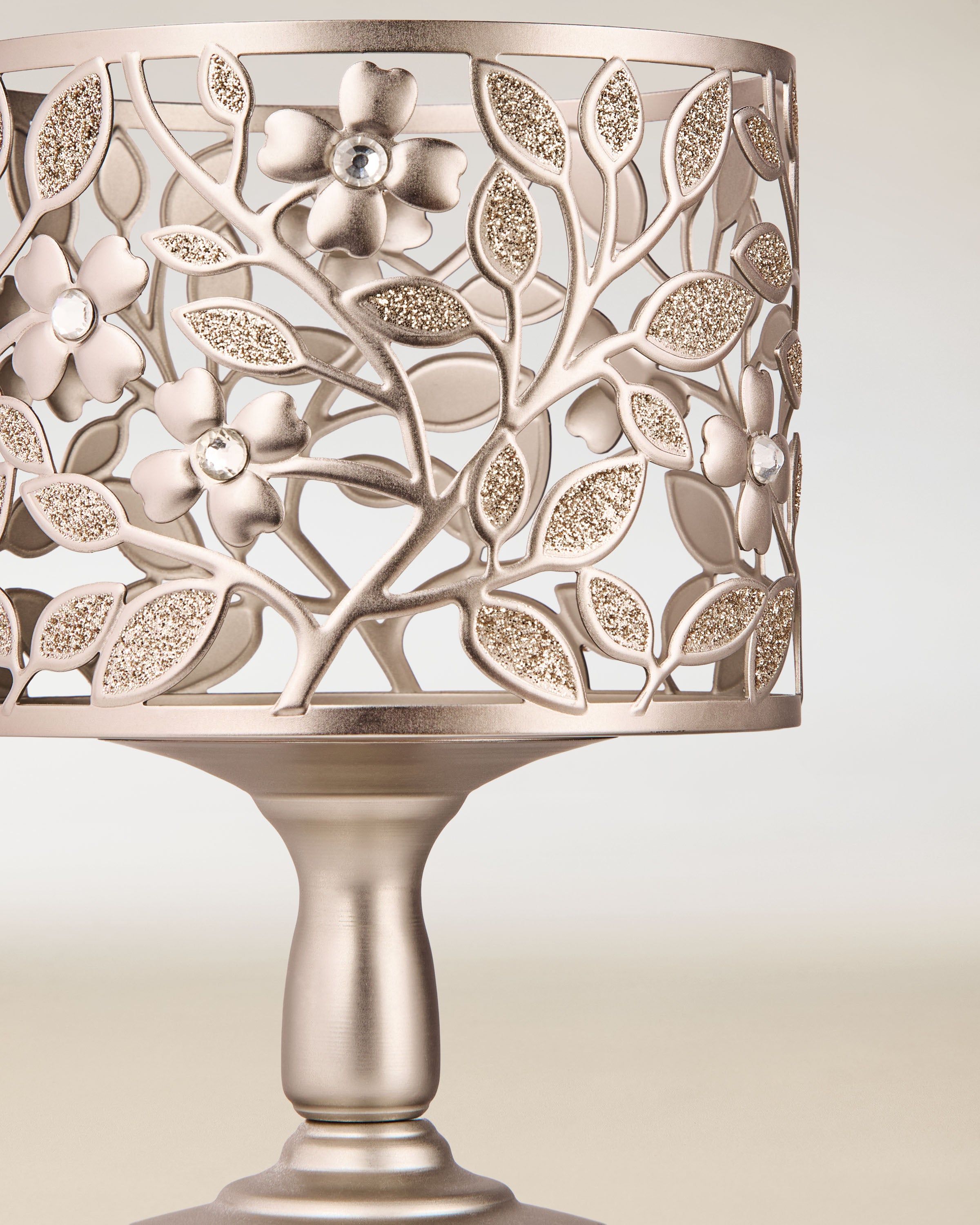 Kovový držák na tříknotové svíčky s jemným květinovým výřezem a třpytivými detaily, umístěný na pevné podstavě.