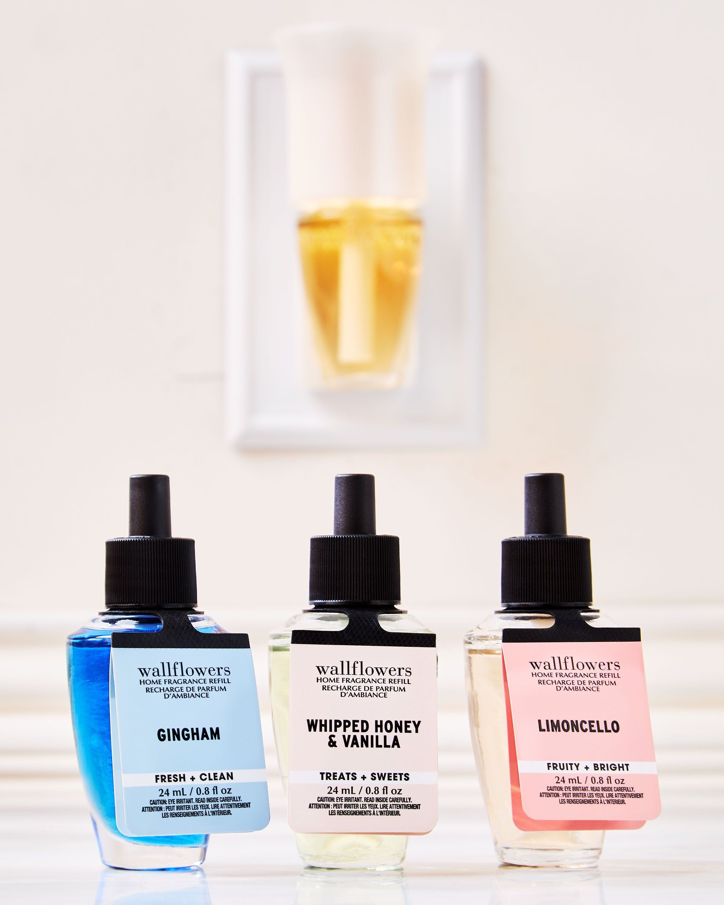 Tři náplně Wallflowers: Gingham, Whipped Honey & Vanilla a Limoncello před nástěnným difuzérem.